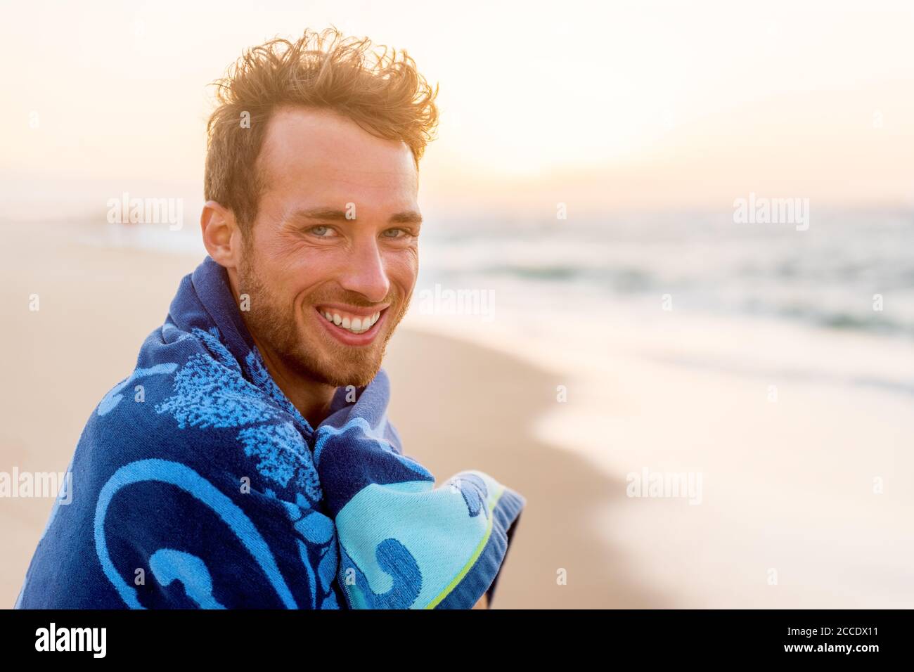 Lächelnd schöner junger Mann Schönheit Porträt am Strand bei Sonnenuntergang Blick auf Kamera lachen, gesundes Grinsen Gesicht glückliches Modell in Handtuch. Stockfoto