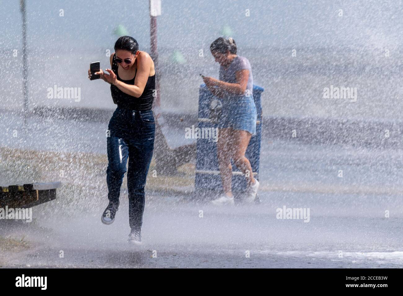 Zwei junge weiße Weibchen, die während der starken Winde des Sturms Ellen in Southend on Sea, Essex, Großbritannien, durch krachende Wellen Rennen, werden nass und durchnässt Stockfoto