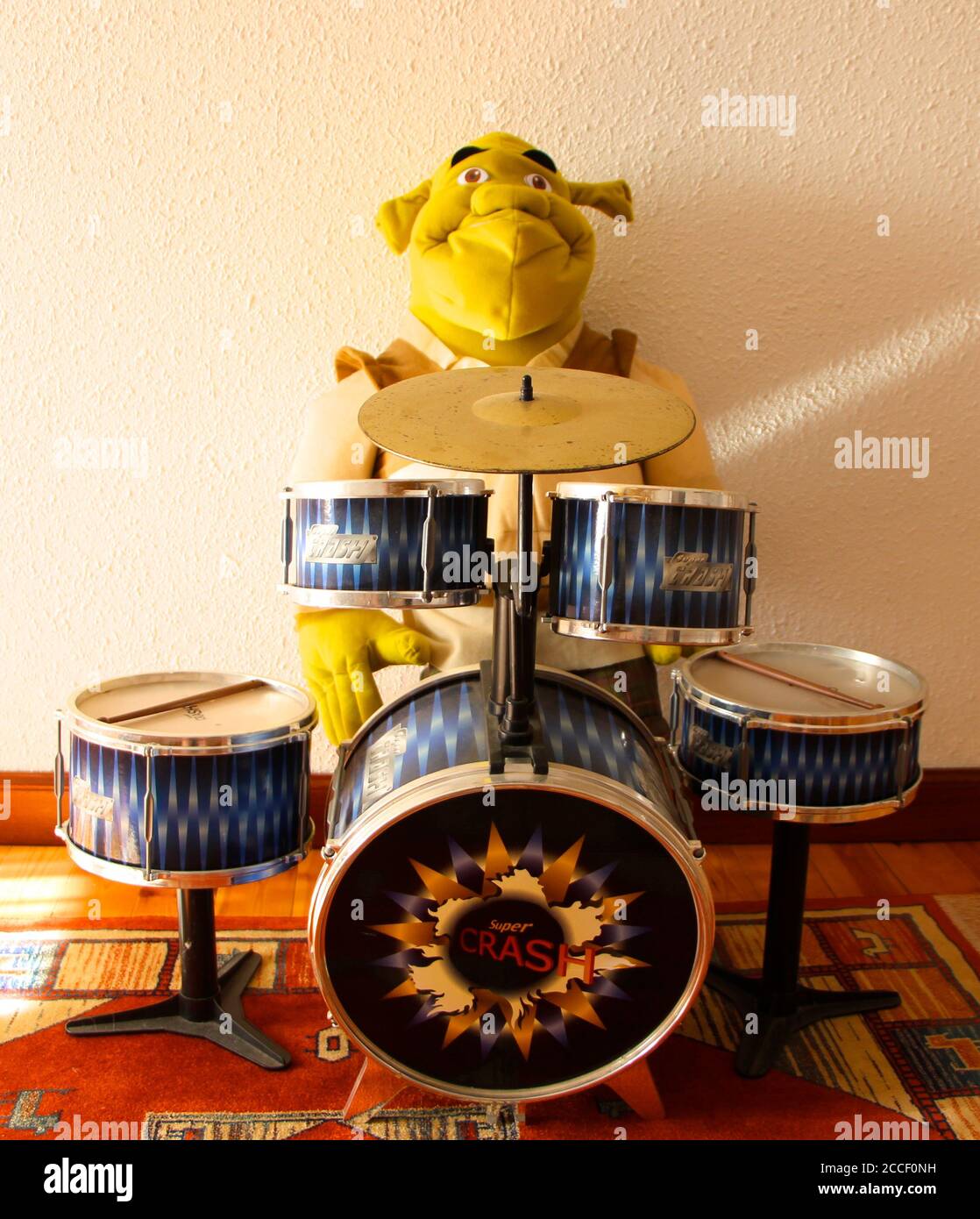 Foto einer großen, weichen Spielzeugfigur der Filmfigur Shrek, die an einem Kinderdrum-Set mit einer weißen Wand und einem roten Teppich am Holzboden sitzt Stockfoto