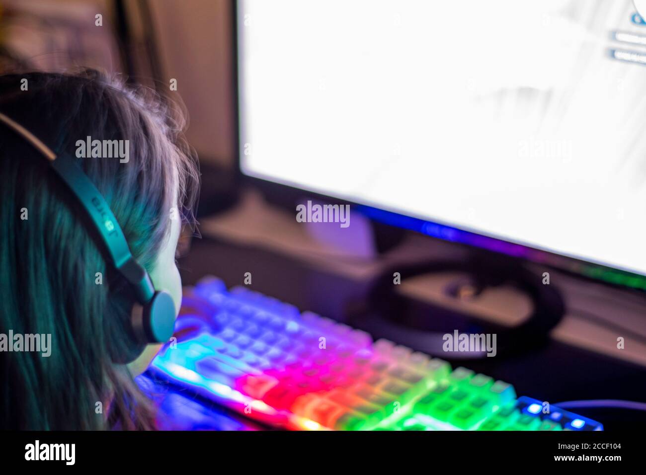 Mädchen der Gaming-Generation mit Headset und farbenfroher Tastatur, die beim Laden von Spielmonitoren Spiele laden möchten. Modernes Lernen. Stockfoto