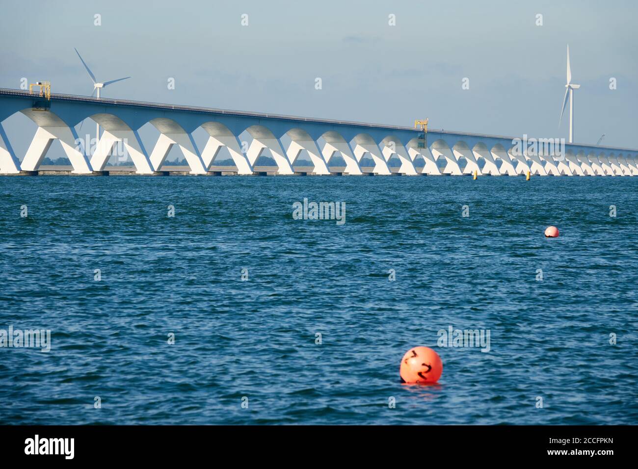 Lange Zeeland Brücke, tiefblaues Wasser mit roter Boje, Windturbine im Hintergrund. Niederlande, Zeeland. Stockfoto