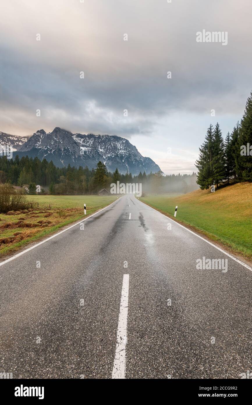 Am frühen Morgen auf der Landstraße am Schmalensee bei Mittenwald ist die Straße von Regen durchnässt, leichter Nebel steigt auf und das Karwendel ragt im Hintergrund auf Stockfoto