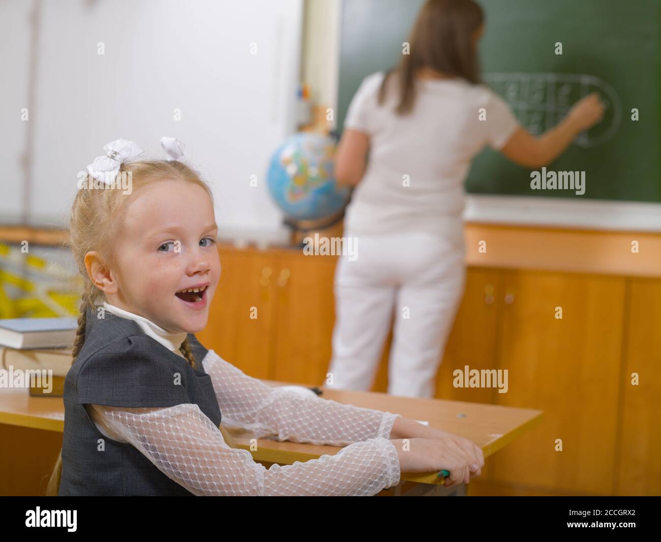 Schulmädchen sitzt am Schreibtisch und dreht sich zurück. Der Lehrer schreibt auf der Tafel im Hintergrund Stockfoto