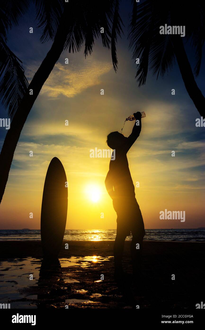 Silhouette von Surf Mann stehen mit einem Surfbrett und Kokosnuss Palm Wasser aus Flasche trinken. Surfen am Strand bei Sonnenuntergang. Outdoor Wassersport Abenteuer Lifesty Stockfoto