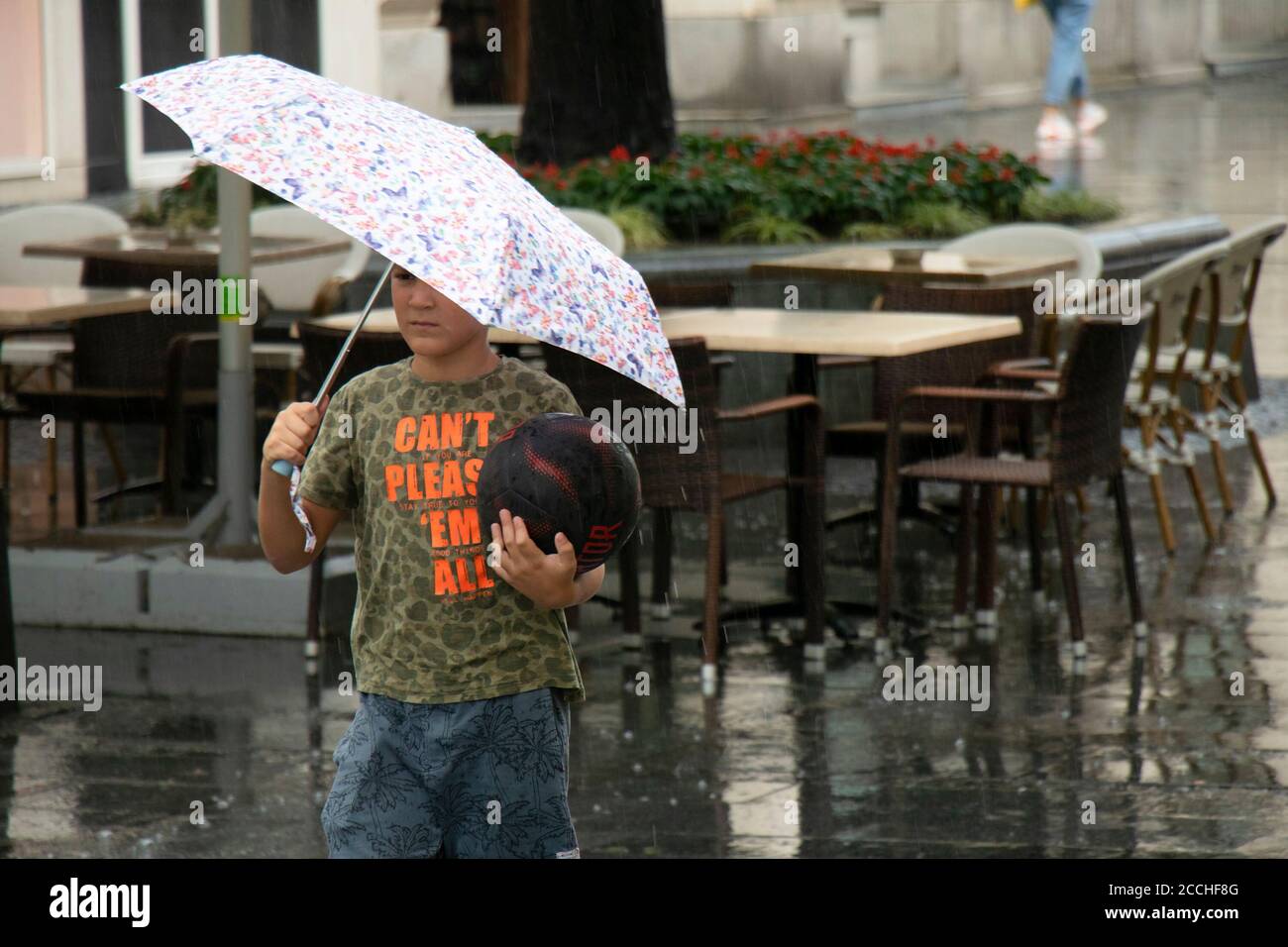 Belgrad, Serbien - 5. August 2020: Kleiner Junge mit einem Basketball, der an einem regnerischen Tag in der Fußgängerzone der Stadt unter dem Regenschirm läuft Stockfoto