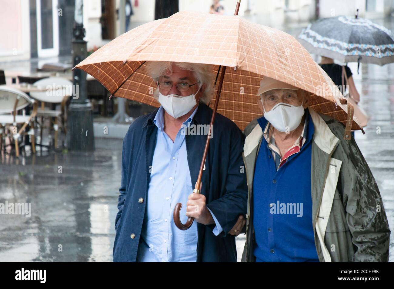 Belgrad, Serbien - 5. August 2020: Zwei Generationen ältere Männer, die eine Schutzmaske tragen, gehen an einem regnerischen Tag auf der Straße unter dem Regenschirm Stockfoto