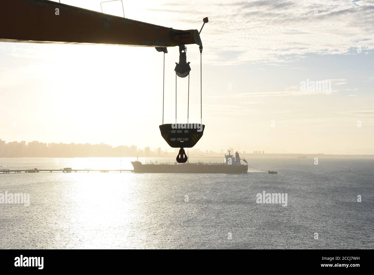 Blick auf die Silhouette eines Tankschiffes unter der Sonne entlang des Entladerohres, das scheinbar am Ausleger und Haken des Frachtschiffes hängt. Stockfoto