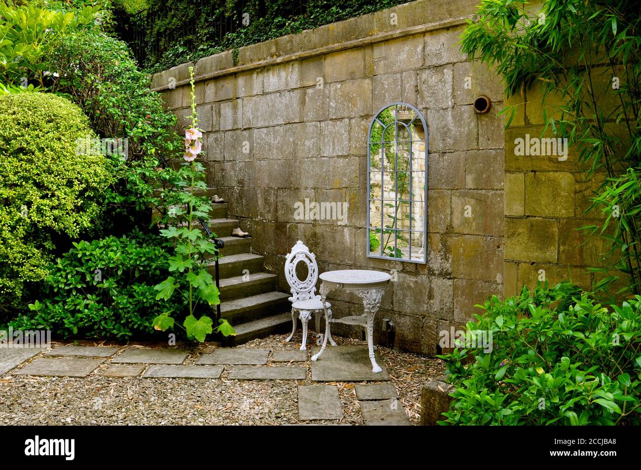 Ein Hof Landgarten mit Steinmauern, Stufen und markierten Wegen. Mit weißen schmiedeeisernen Terrassenmöbeln, einem Spiegel im georgianischen Stil und üppigen Bäumen, Stockfoto