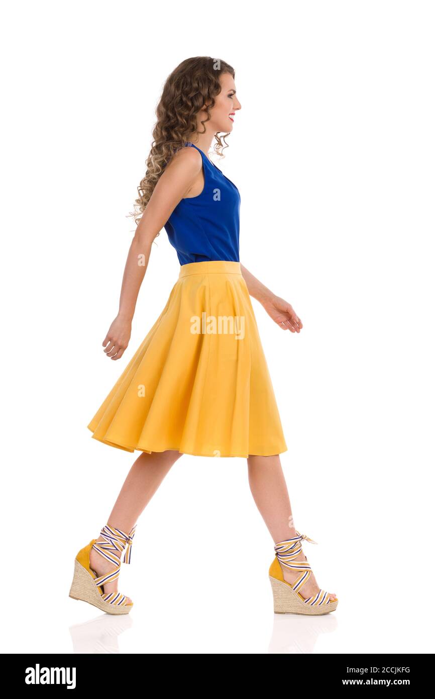 Schöne junge Frau in gelbem Rock, blauem Top und Keilschuhe geht und schaut weg. Seitenansicht. Studioaufnahme in voller Länge, isoliert auf Weiß. Stockfoto