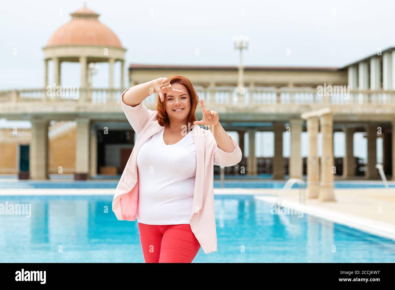 Eine glückliche kaukasische Plus-Größe Frau hält einen Rahmen für ein Foto-Shooting. Ein Park mit Schwimmbad im Hintergrund. Konzept der menschlichen Emotionen, busi Stockfoto