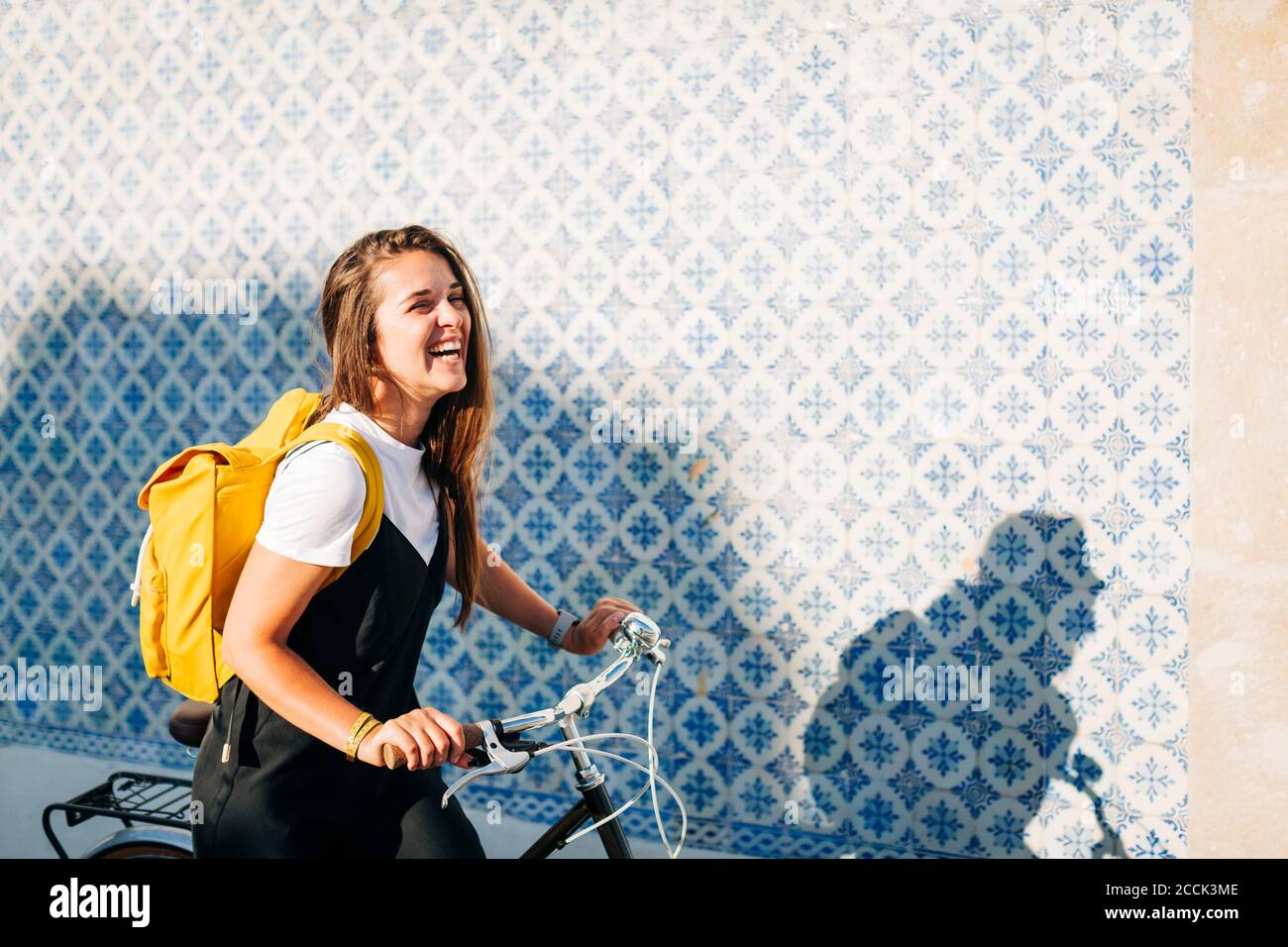 Fröhliche Frau mit Rucksack Fahrrad von gefliesten Wand in reiten Stadt an sonnigen Tag Stockfoto