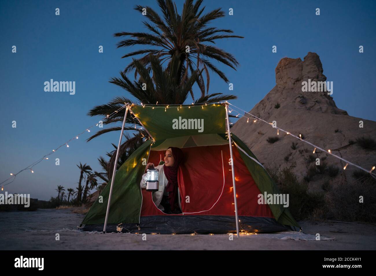 Lächelnde junge Touristenfrau in Hijab, die aus dem Zelt schaut In der Abenddämmerung Stockfoto