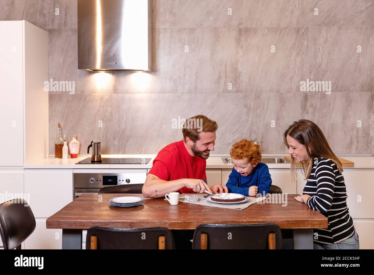 Glücklicher Mann, der Schokoladenkuchen schneidet, während er mit der Familie sitzt Küche Stockfoto