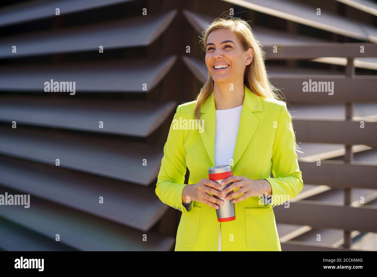 Lächelnde Geschäftsfrau mit Reisetasse, die wegschaut, während sie dagegen steht Geländer Stockfoto