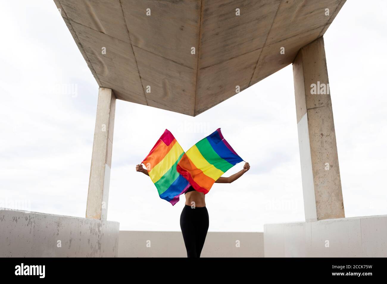 Mittlere Erwachsene Frau winkt Regenbogenfahnen, während sie auf gebaut steht Struktur gegen Himmel Stockfoto