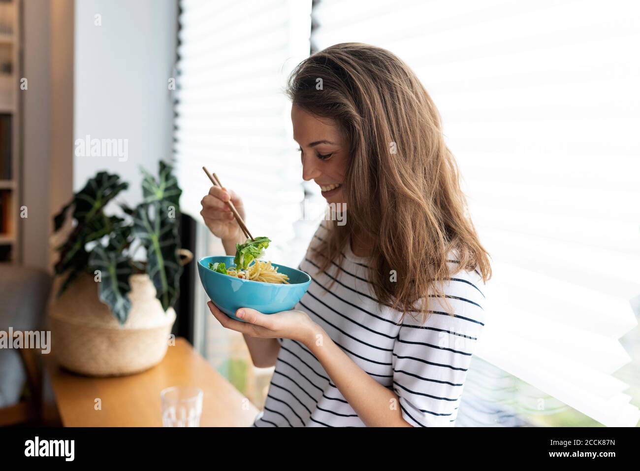 Glückliche junge Frau beim Essen, während sie gegen das Fenster steht Zu Hause Stockfoto