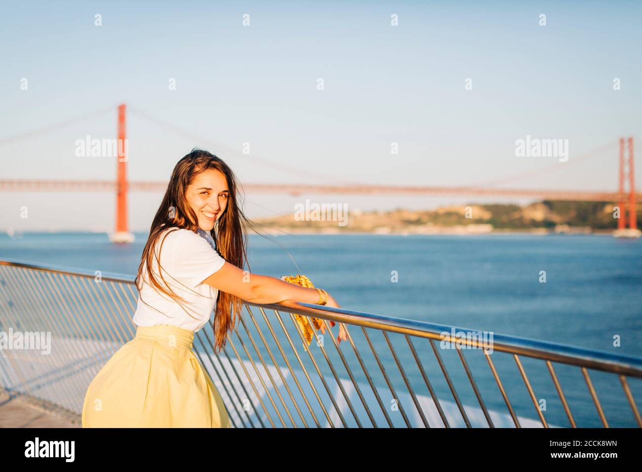 Lächelnd schöne Frau mit langen Haaren, die beim Geländer stehen Fluss in der Stadt bei Sonnenuntergang Stockfoto