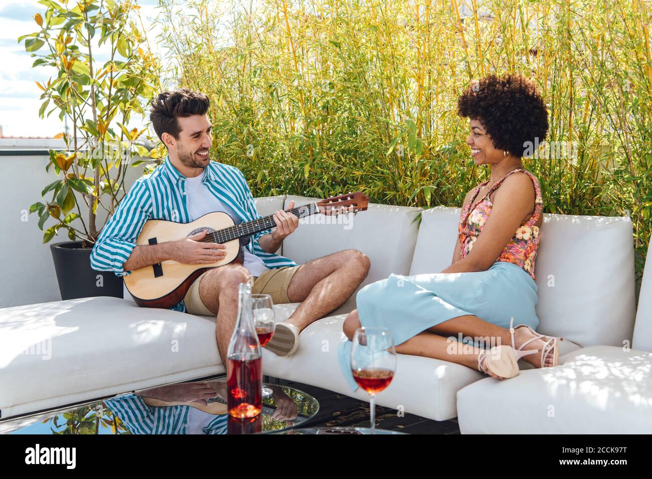 Glücklicher Mann, der Gitarre für Frau spielt, während er auf dem Sofa sitzt Im Penthouse Patio Stockfoto