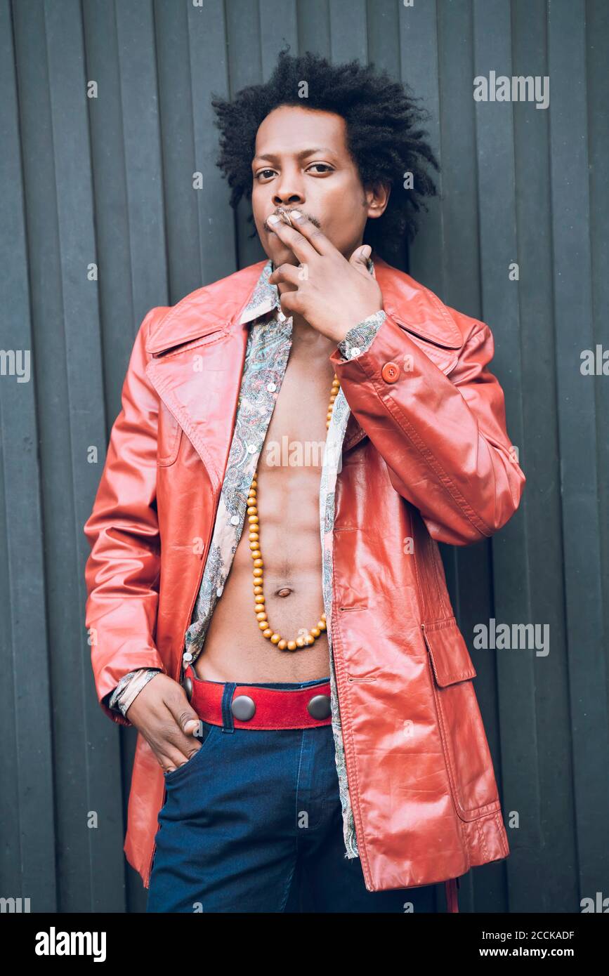 Selbstbewusster Mann in Retro-Jacke rauchen, während sie gegen schwarz stehen Wand Stockfoto