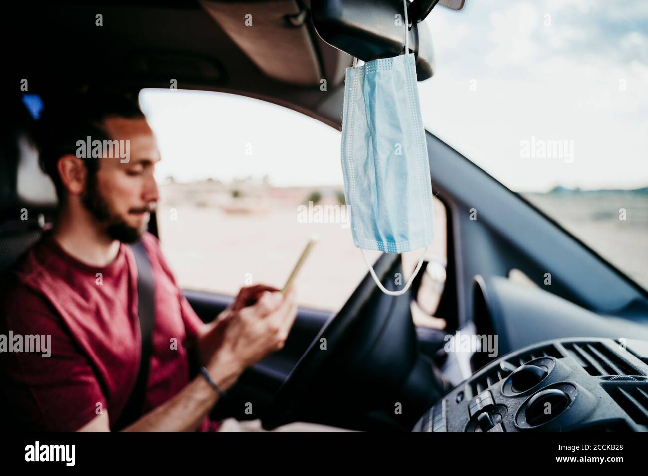 Nahaufnahme der Gesichtsmaske hängt auf Rückspiegel, während Mann Telefon im Auto verwenden Stockfoto