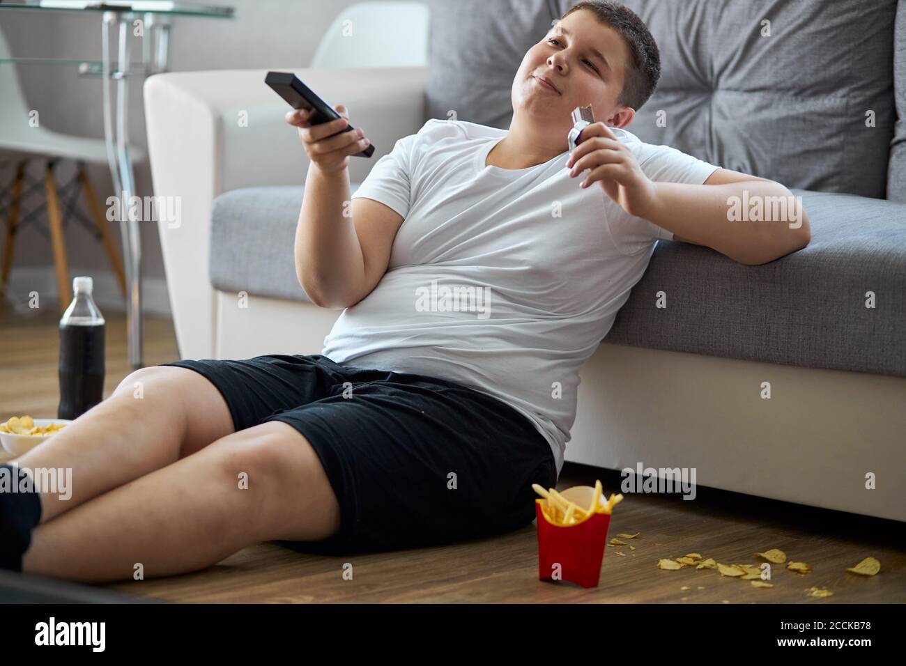 Übergewicht Fett Junge essen Junk-Food beim Fernsehen allein zu Hause, sitzen auf dem Boden mit pommes frites, kohlensäurehaltige Getränke, Chips. Genießen Sie ungesunde Heftigkeit Stockfoto