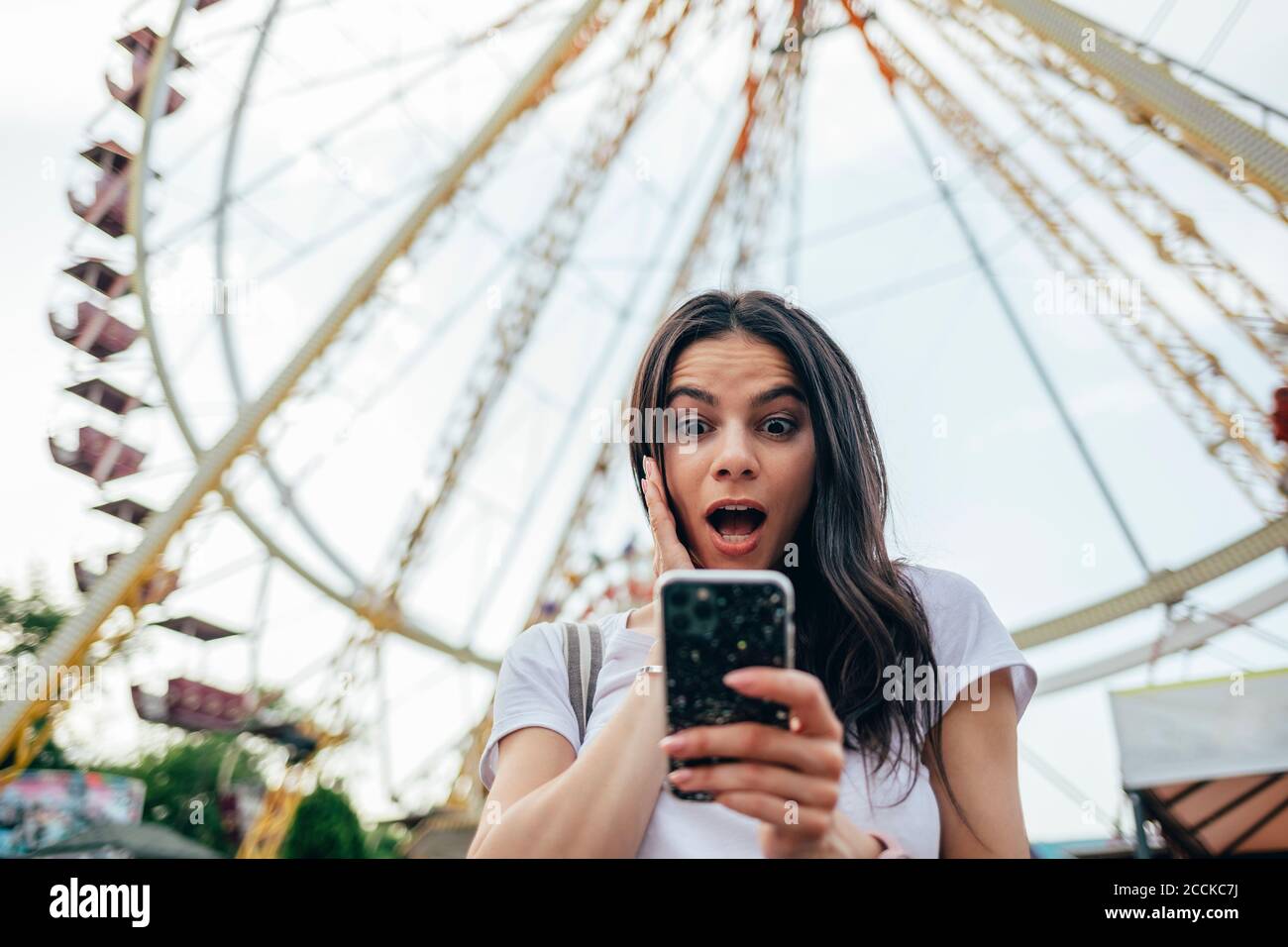 Überrascht junge Frau mit Mund offen mit Smartphone gegen Riesenrad im Vergnügungspark Stockfoto