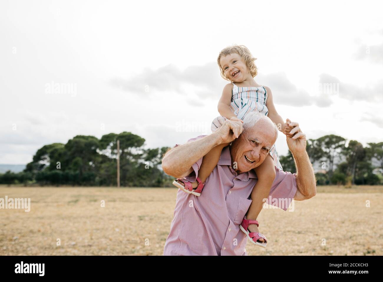 Glücklicher Großvater trägt Enkelin auf Schultern, während er gegen den Himmel steht Stockfoto