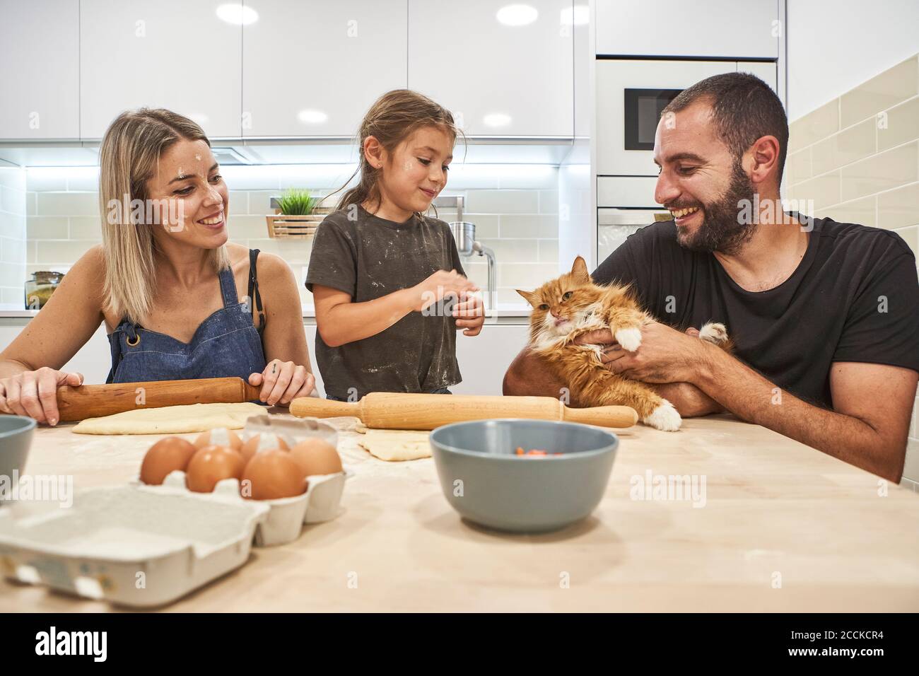 Mutter knetet Teig mit Nudelholz, während Vater und Tochter Spielen mit der Katze auf dem Küchentisch Stockfoto