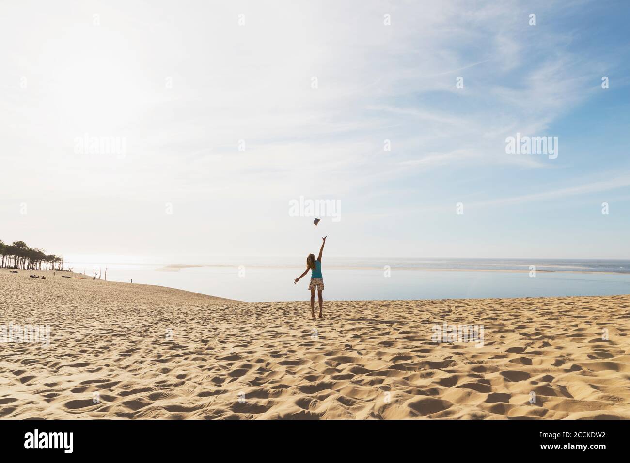 Glückliche Frau, die Hut in der Luft wirft, während sie am Strand gegen den Himmel steht, Düne von Pilat, Nouvelle-Aquitaine, Frankreich Stockfoto