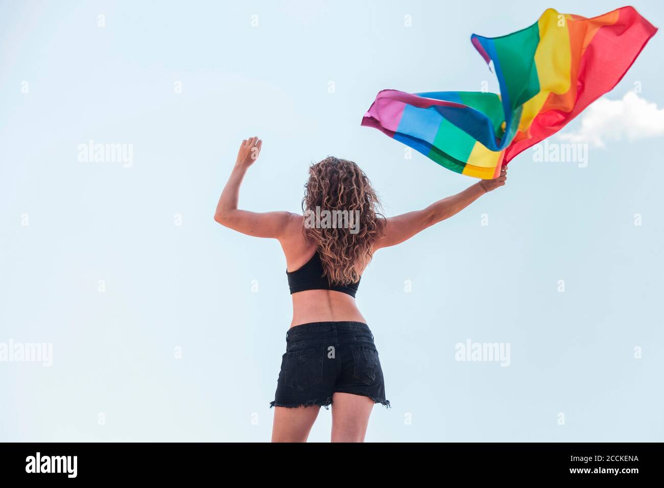 Frau mit mittlerem Erwachsenen, die Regenbogenfahne schwenkt, während sie gegen den Himmel steht Stockfoto