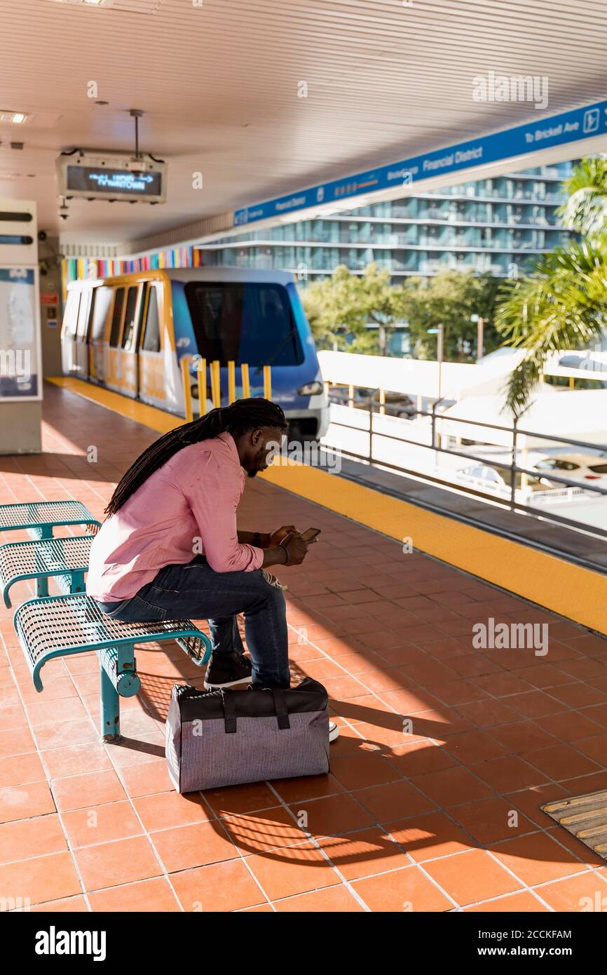 Junger Mann, der Smartphone benutzt, während er auf dem Sitz am Bahnhof, Miami, Florida, USA sitzt Stockfoto