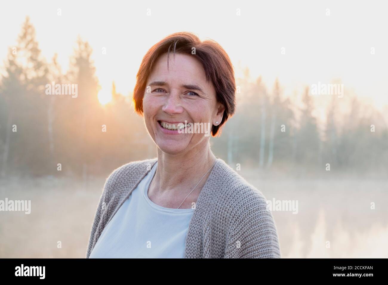 Porträt einer erwachsenen Frau, die während des nebligen Sonnenaufgangs an der Kamera lächelt Stockfoto