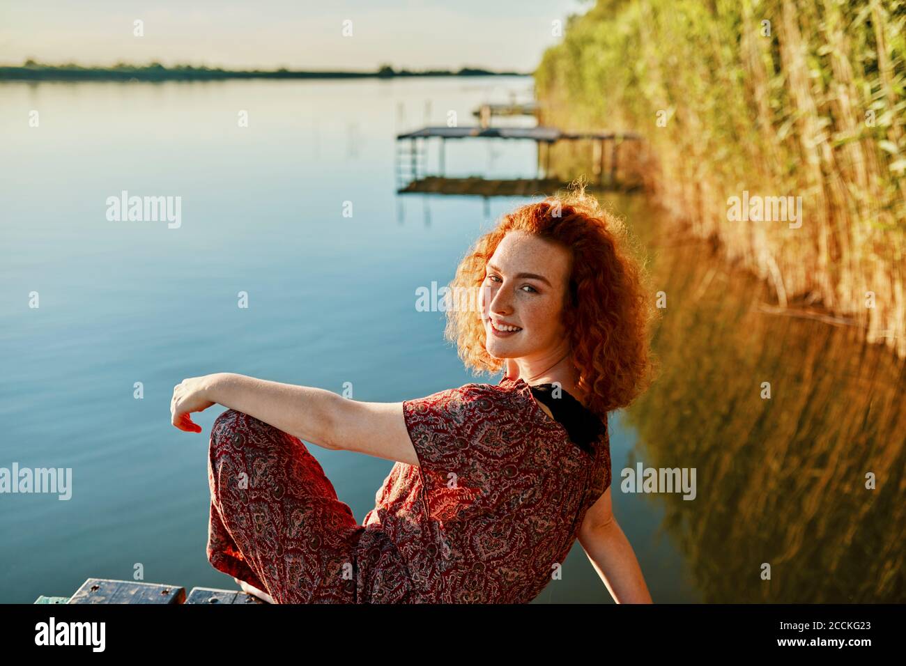 Porträt einer lächelnden rothaarigen jungen Frau, die auf dem Anlegesteg sitzt Ein See bei Sonnenuntergang Stockfoto