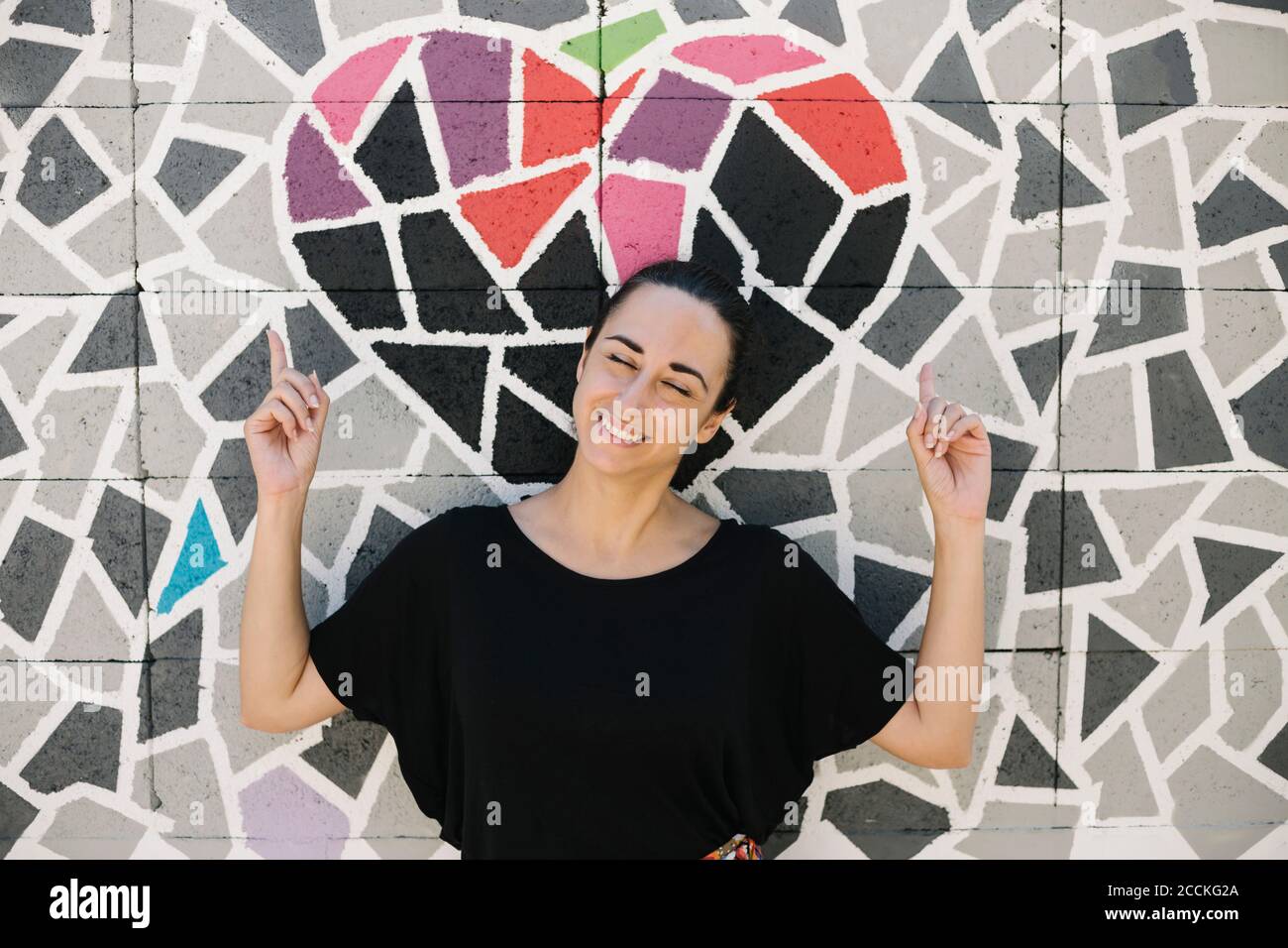 Portrait der lächelnden Frau zeigt auf Herz auf bunten Wand Mosaik Stockfoto