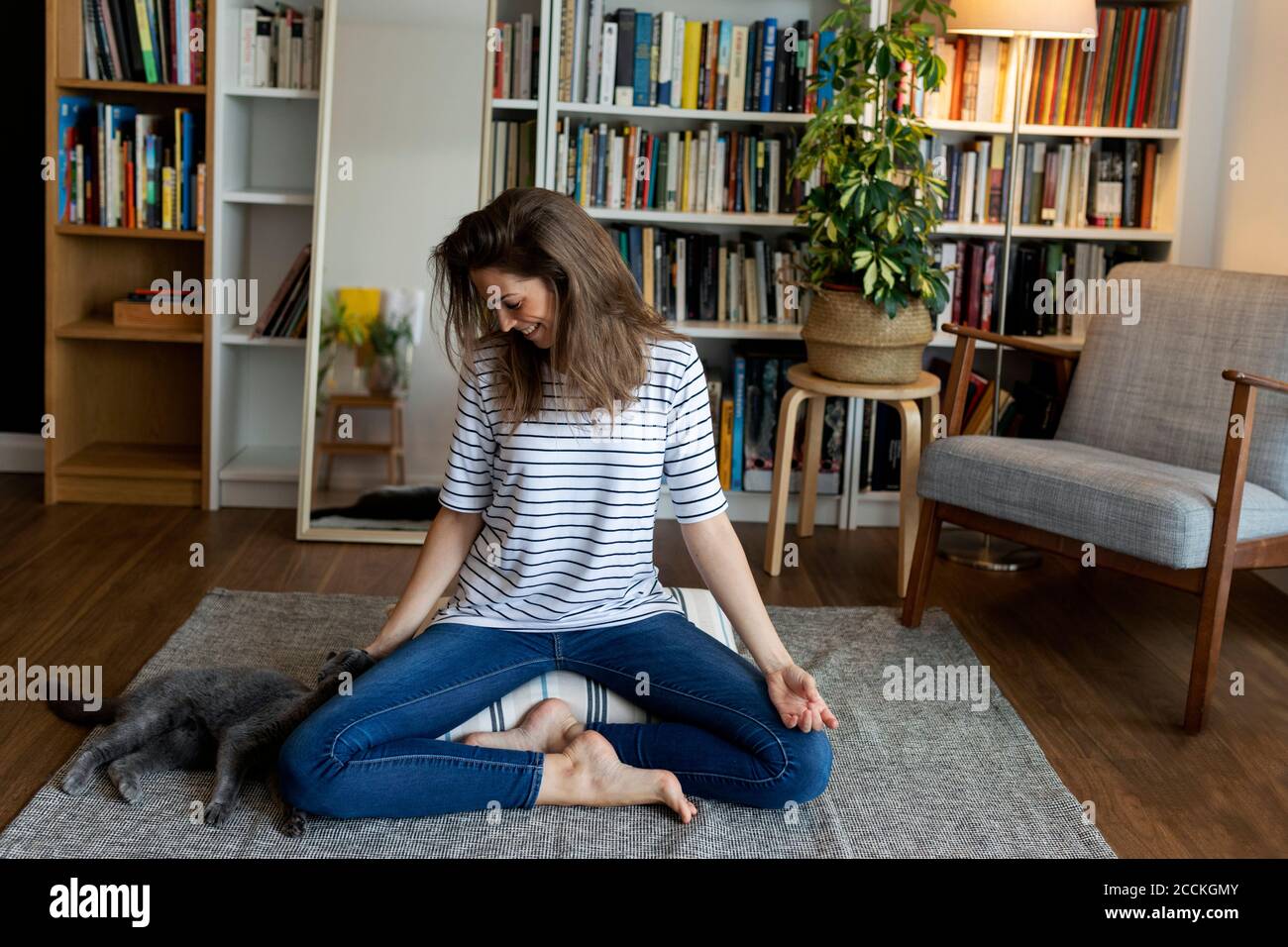 Glückliche junge Frau mit Katze sitzt auf dem Teppich gegen Bücherregal Zu Hause Stockfoto