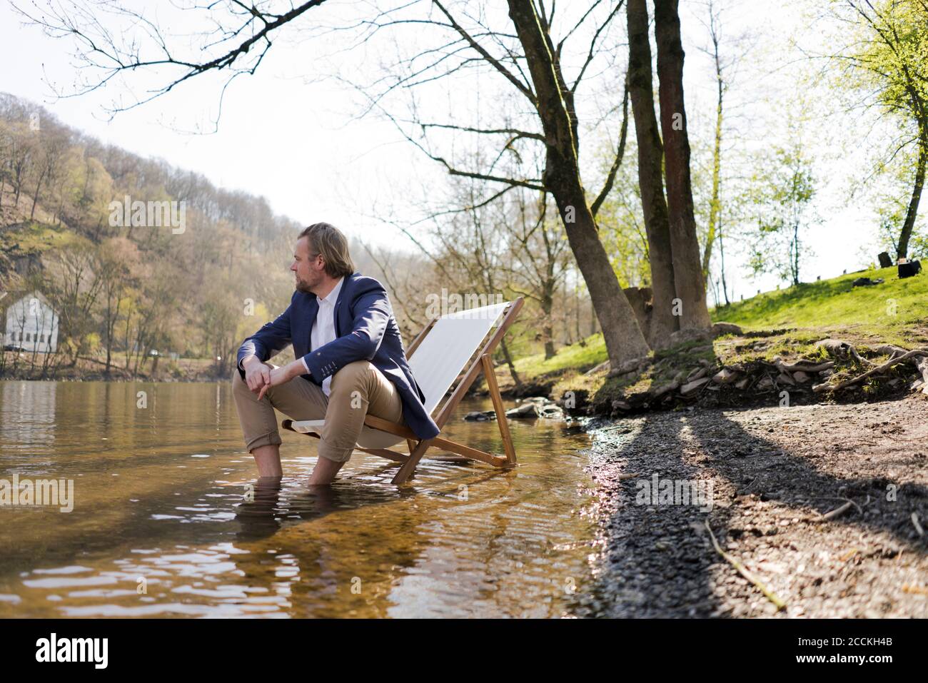 Nachdenklich männlichen professionellen Blick weg, während auf Stuhl sitzen Flussufer gegen Bäume im Park Stockfoto