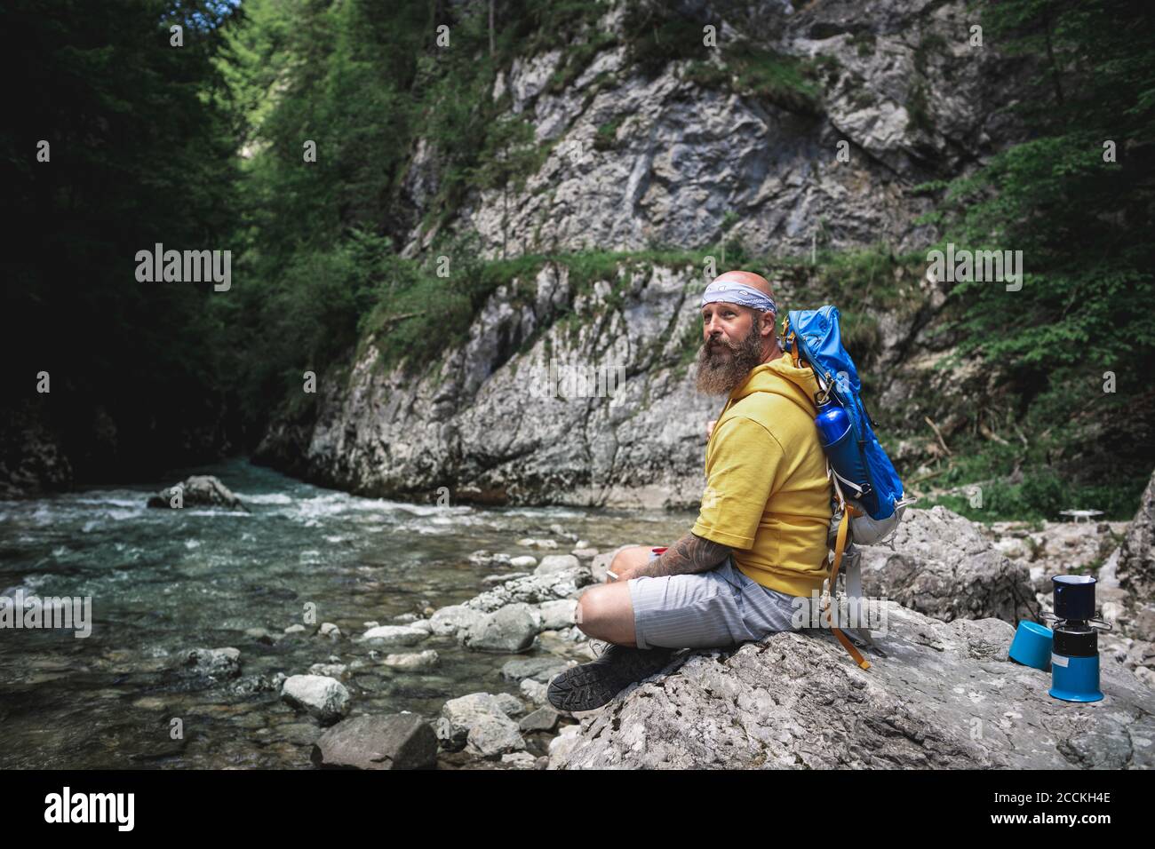 Wanderer mit Vollbart und gelbem Hoodie auf Stein sitzend Neben dem Fluss Stockfoto