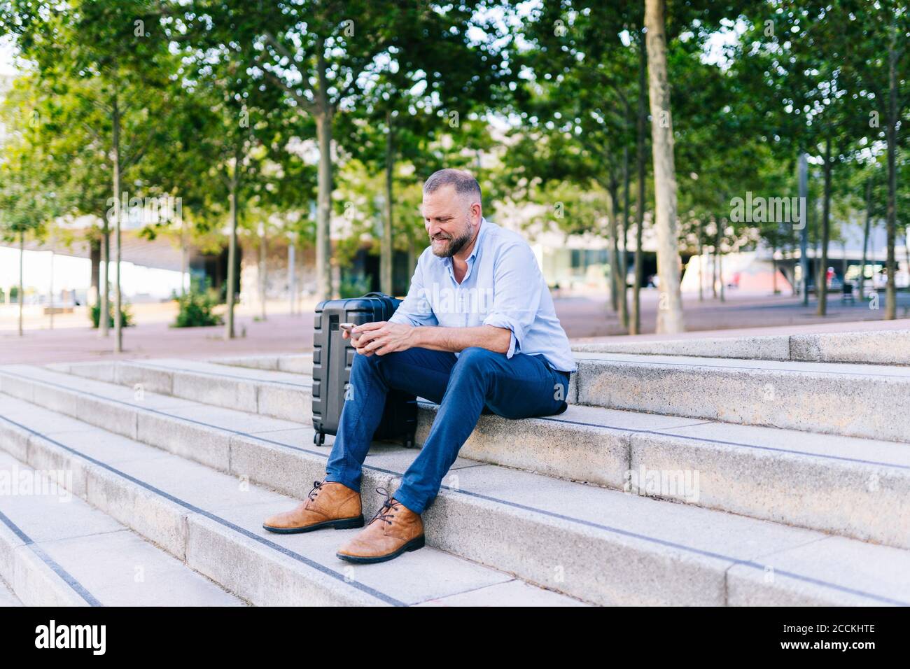 Geschäftsmann mit Handy, während er am Koffer auf der Treppe sitzt Stockfoto