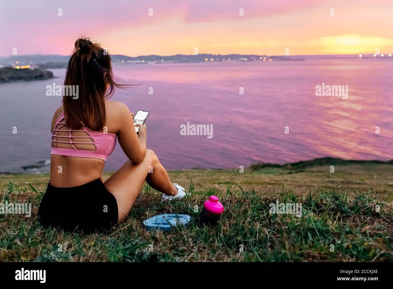 Sportlerin mit Smartphone auf Gras sitzend, während sie anschaut Meer gegen Himmel bei Sonnenuntergang Stockfoto