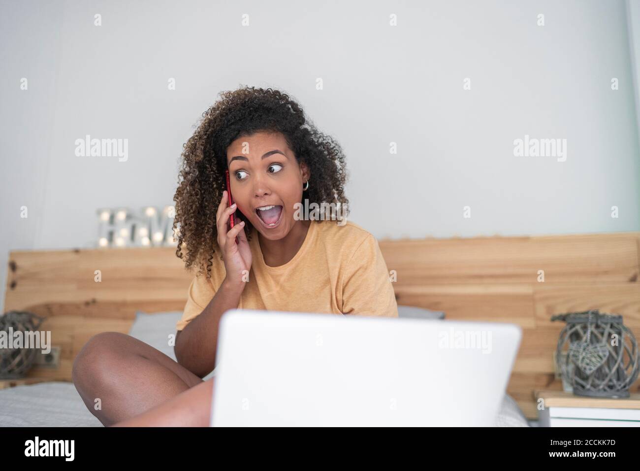 Fröhliche Frau mit offenem Mund reden über Smartphone, während Sitzen auf dem Bett zu Hause Stockfoto