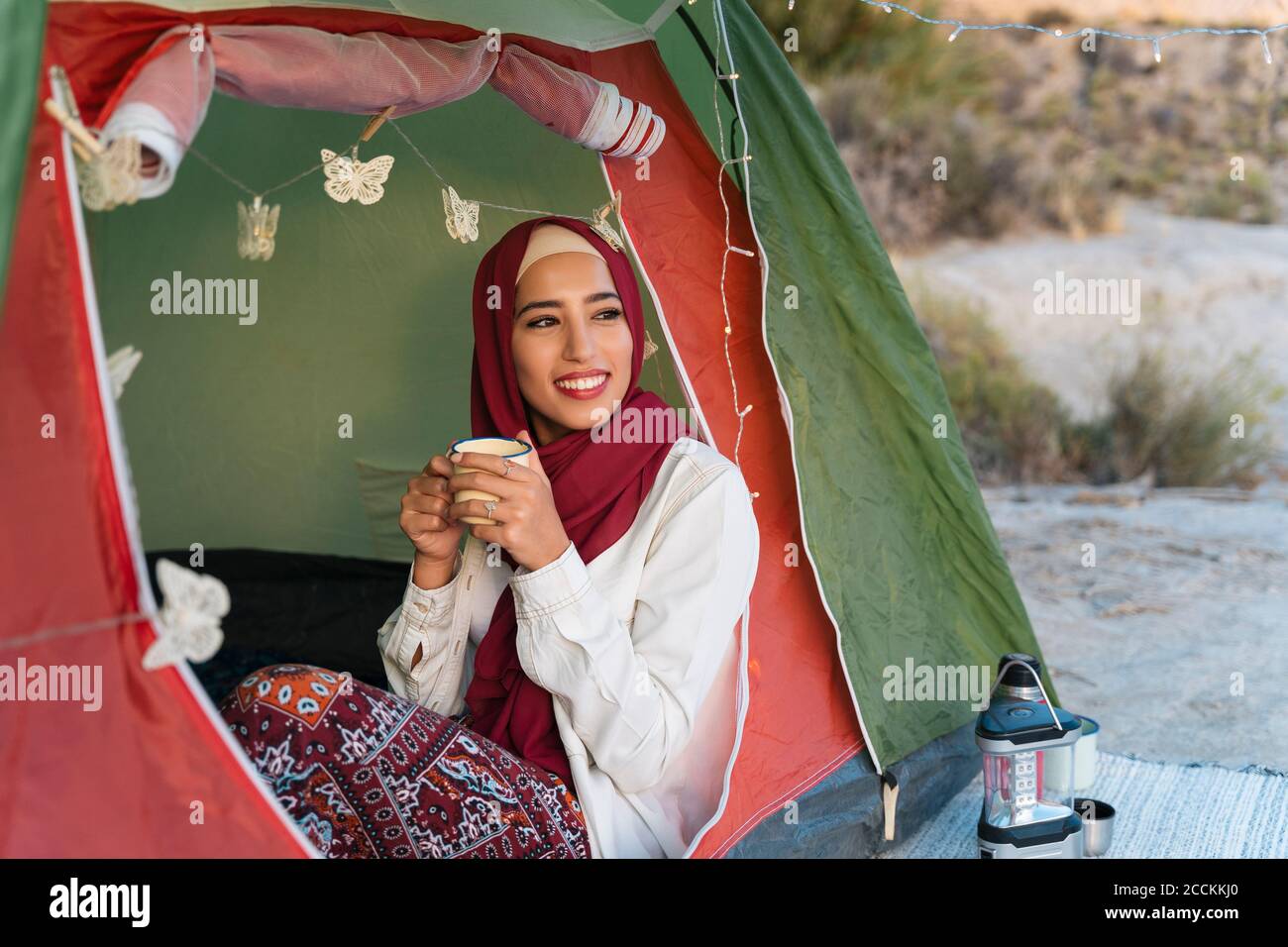 Lächelnde junge Touristenfrau trägt Hijab in einem Zelt halten Ein Becher Stockfoto
