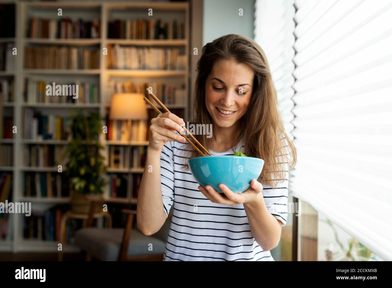 Lächelnde junge Frau, die beim Essen am Fenster sitzt Zu Hause Stockfoto