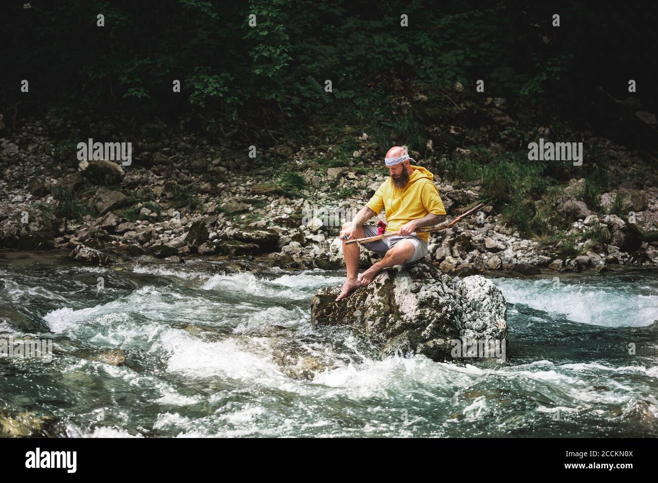 Abenteurer mit Bart sitzt in der Mitte des Flusses auf Stein und Schnitzwerk auf einem Stück Holz Stockfoto