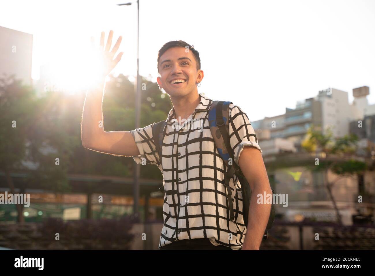 Lächelnder junger schwuler Mann winkt, während er in der Stadt gegen steht Himmel Stockfoto