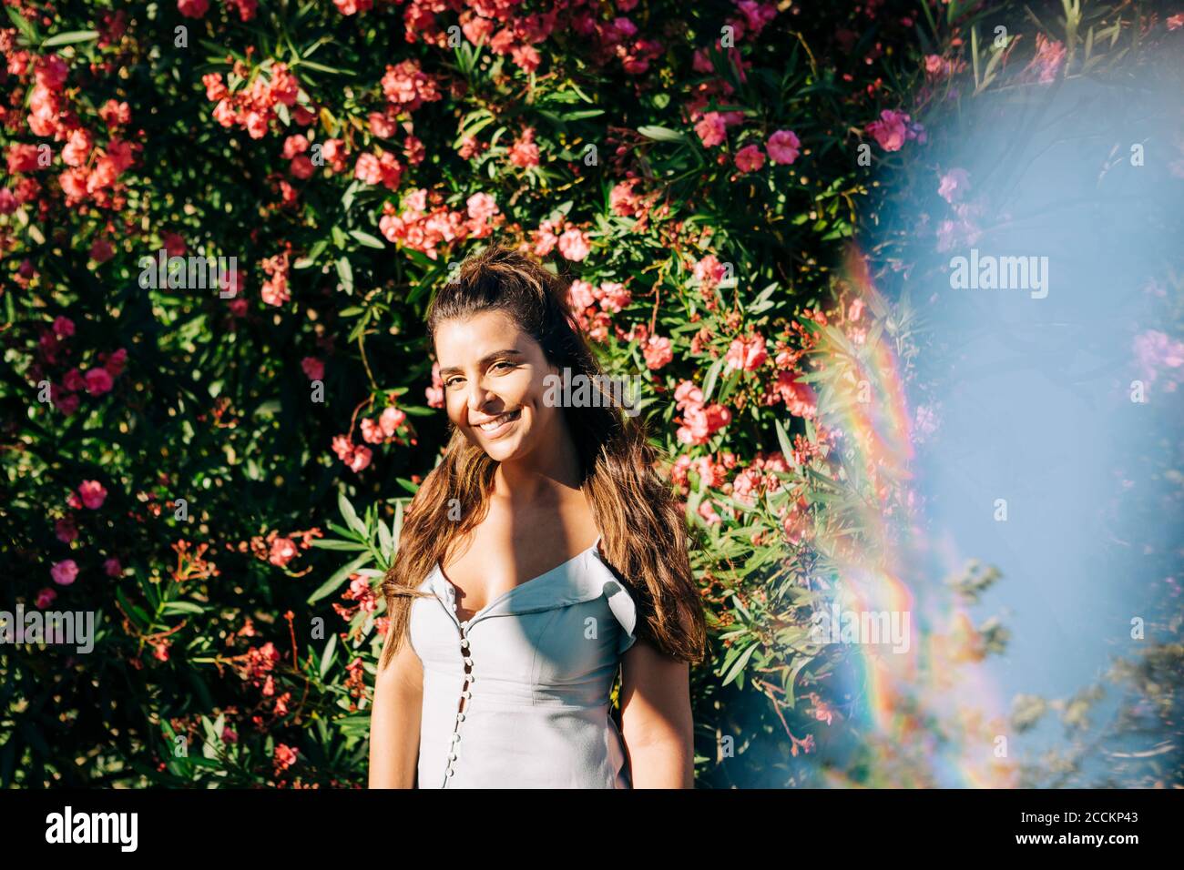 Lächelnde junge Frau mit langen Haaren, die gegen Pflanzen stehen park an sonnigen Tag Stockfoto