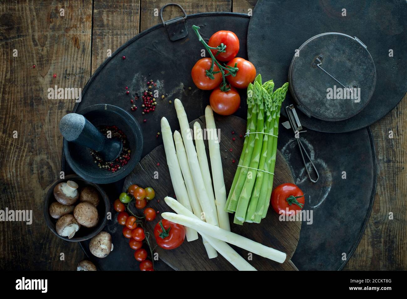 Mörtel und Stössel, Schäler, Spargelstiele, Pfefferkorn, Tomaten und Schüssel mit Pilzen auf rustikalem Backblech Stockfoto