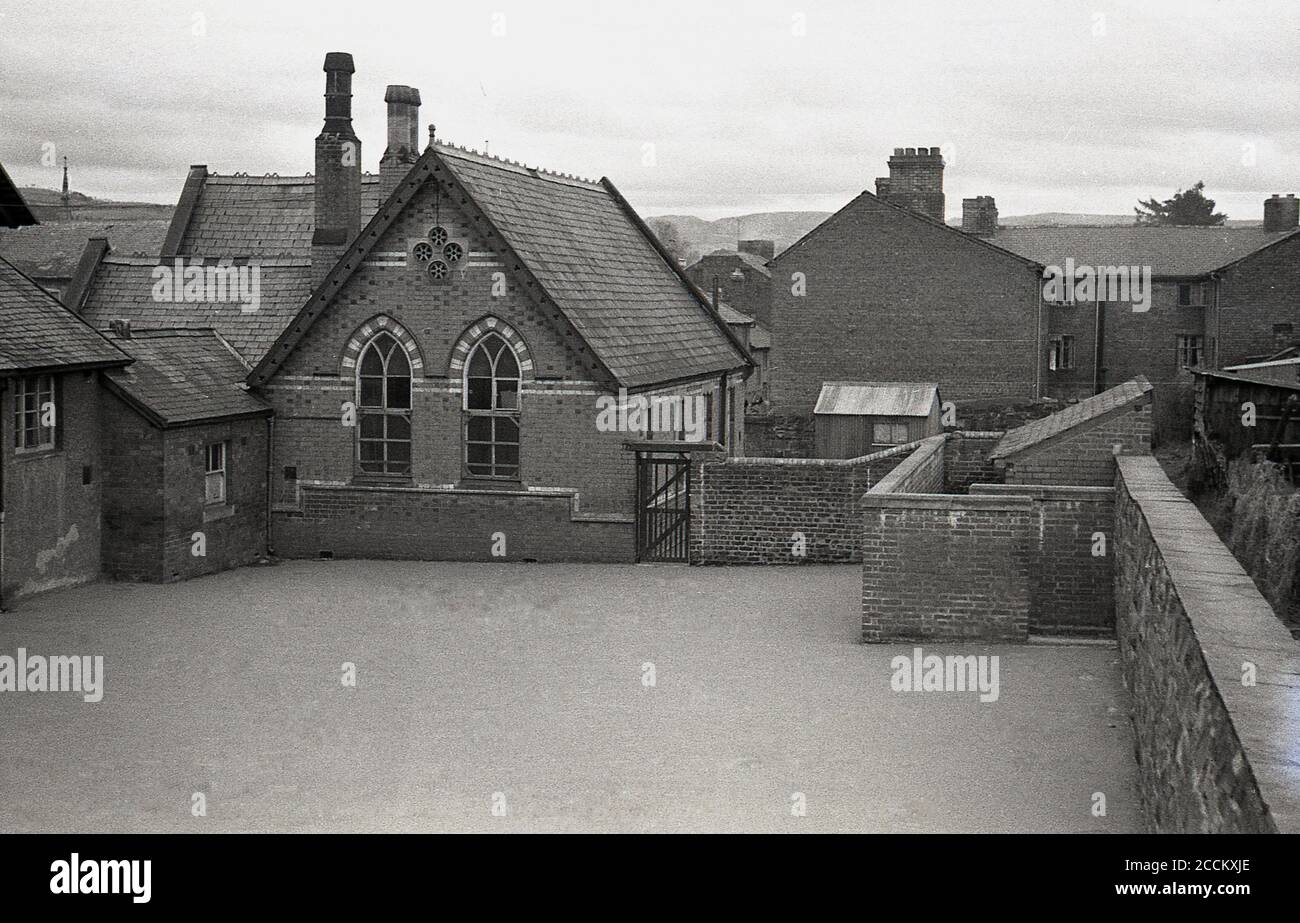 1950er Jahre, historisches Bild aus dieser Zeit zeigt die Dorfschule, Gebäude und geschlossenen Spielplatz, Llandyssil, Powys, Wales. Die viktorianische Schule wurde 1866 gebaut, entworfen von Thomas Henry Wyatt, einem anglo-irischen Architekten, der eine herausragende Karriere hatte, wurde 1870 zum Präsidenten des Royal Institute of Briitsh Architects gewählt und erhielt 1873 die Royal Gold Medal for Architecture. Die Schule schloss 1950 und Teile wurden erst 2010 durch eine Spendenaktion gerettet, um die Schulhalle zu einem lokalen Gemeindezentrum zu machen. Stockfoto