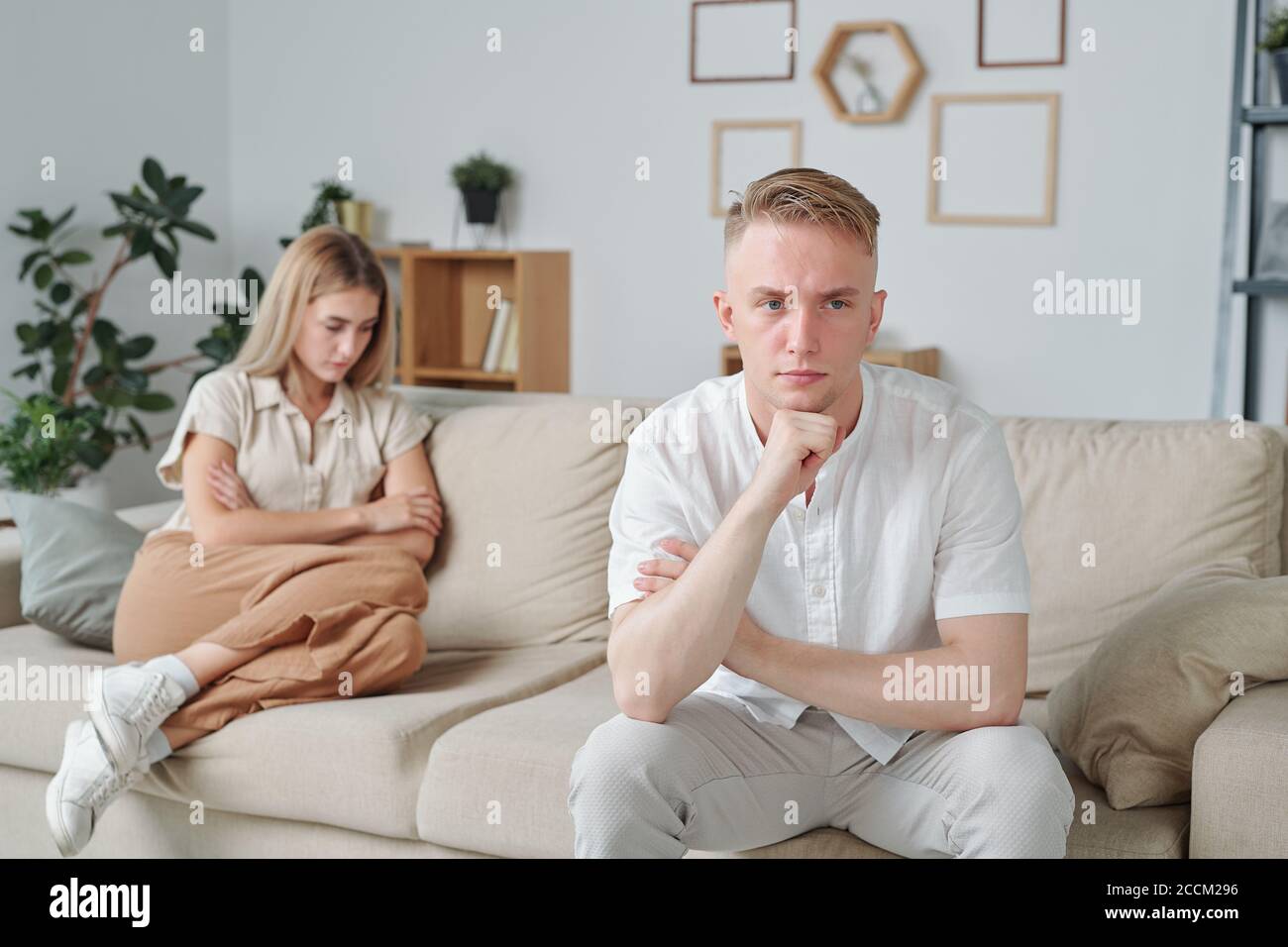 Junger beleidigter Mann sitzt auf der Couch auf dem Hintergrund seiner Unglückliche Frau Stockfoto