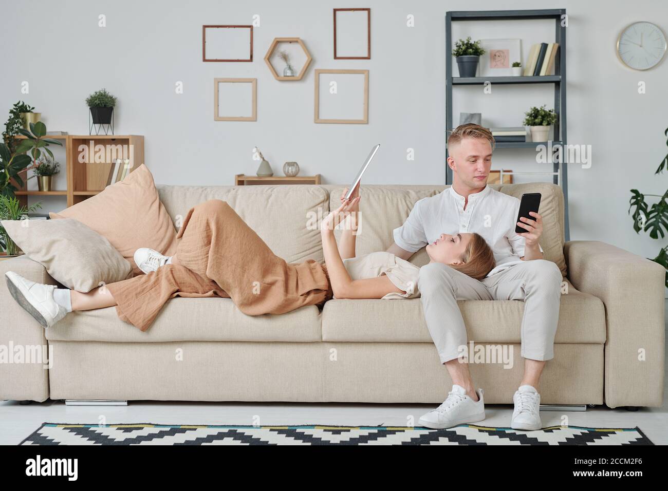 Junge Frau mit Touchpad hält ihren Kopf auf den Knien Ehemann mit Smartphone Stockfoto