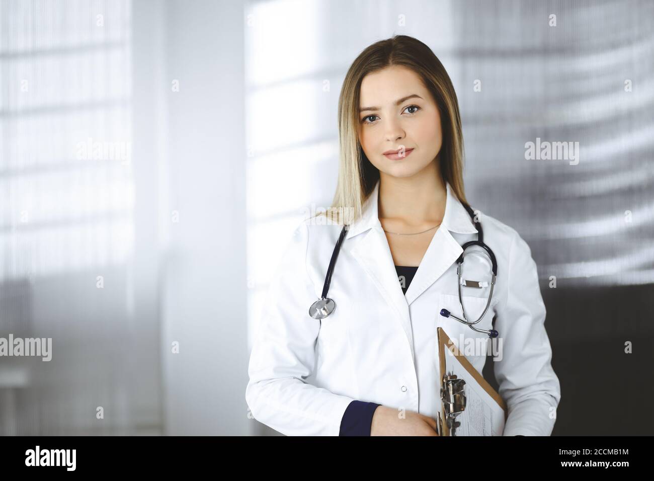 Junge optimistische Ärztin hält ein Klemmbrett in den Händen, während sie in einer Klinik steht. Porträt einer freundlichen Ärztin mit einem Stockfoto